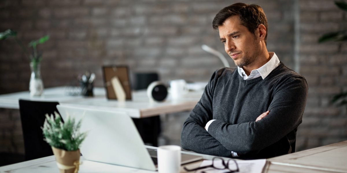 Displeased businessman with arms crossed reading an e-mail on laptop at work.