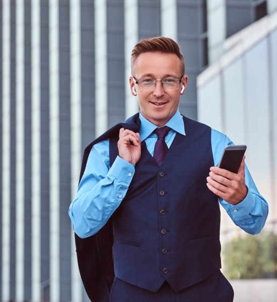 Smiling businessman dressed in an elegant suit holds smartphone and jacket on the shoulder while standing against cityscape background.