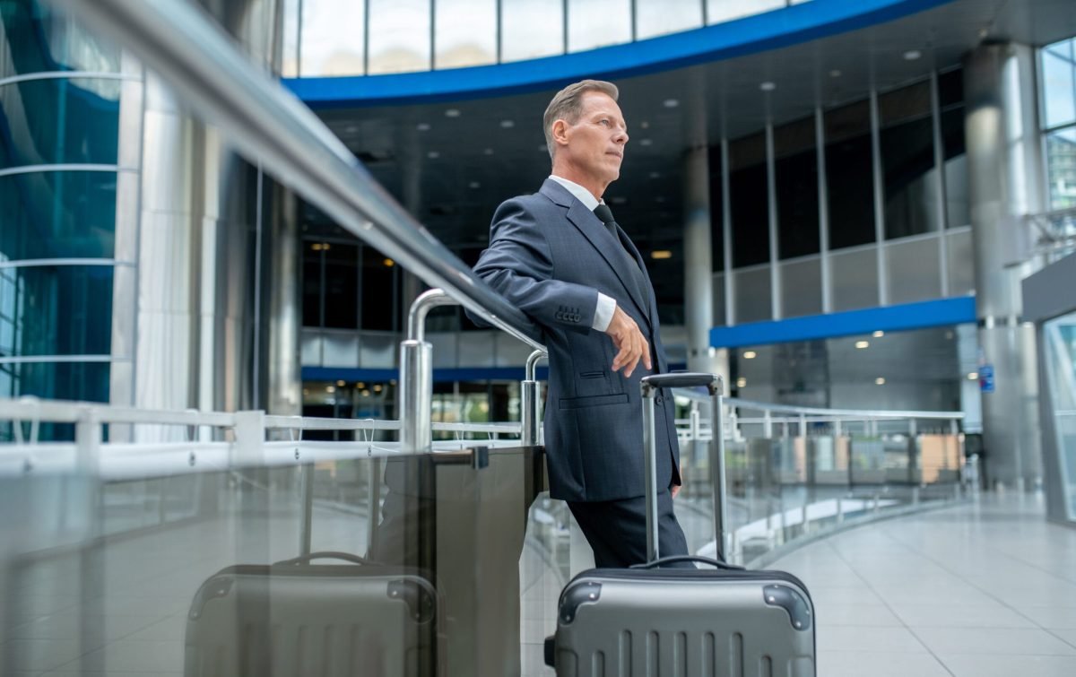 Tranquil pensive businessman with the suitcase standing at airport terminal Calm thoughtful male entrepreneur with his baggage leaned on the handrail staring into the distance